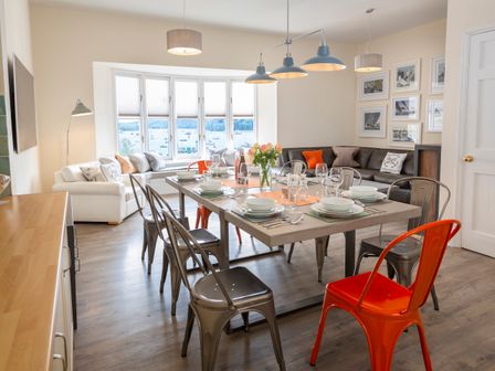 A dining table set with dishes and glasses surrounded by metal chairs in a living room with sofas and a bay window at Coxswains Watch in Salcombe
