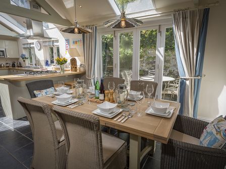 A dining area with six wicker chairs around a wooden table set with dishes and glasses next to glass doors with curtains and an adjacent kitchen at Gooder House in Dartmouth