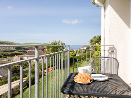 A balcony with a black metal table and chair and pastries and mugs with a view of houses greenery and the sea at Grand View in Hope Cove