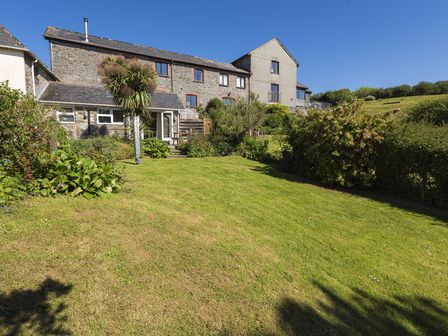 A backyard with green grass shrubs and a stone house under a clear blue sky at Hazel Cottage in Malborough