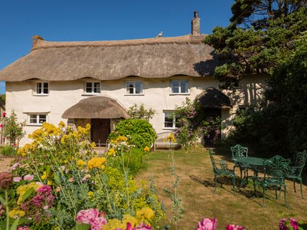 A garden with flowers and green metal table and chairs in front of a thatched cottage at Higher Collaton Cottage in Malborough