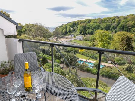 A balcony with a metal table and four glasses and a bottle overlooking a garden with a swimming pool and trees at The Hoot 16 St Elmo Court in Salcombe