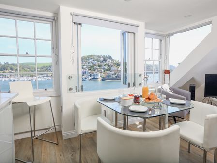 A dining area with a glass table set for breakfast surrounded by white chairs and large windows with a view of water and hillside at Kings View in Dartmouth