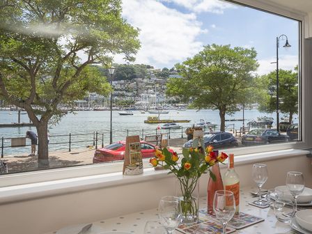 A dining table set with wine bottles glasses and flowers by a window with a view of a waterfront street with cars trees and boats at Leeside in Dartmouth