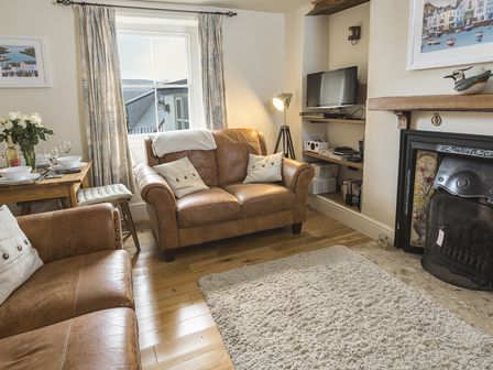A living room with brown leather sofas a dining table with tableware a window with curtains a floor lamp a television a fireplace and a rug at Pixie Cottage in Dartmouth