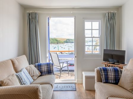 A living room with two beige sofas facing each other and a chair on a balcony overlooking a harbor at Spinnaker Cottage in Salcombe
