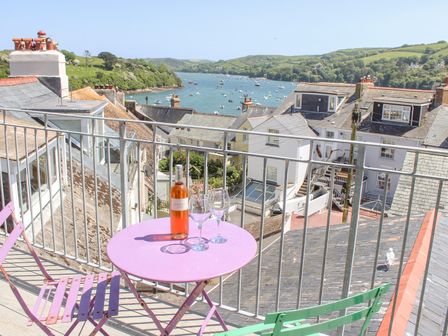 A balcony with a pink table and two chairs with a bottle of wine and two glasses overlooking rooftops and a river with boats at Upper Marcam House in Salcombe
