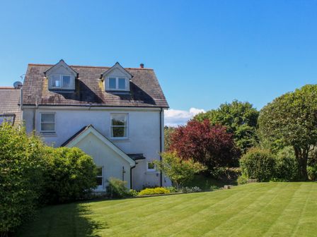 A white house with a steep roof and dormer windows next to a lawn and trees at Warren Cottage in Dartmouth