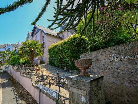 The exterior of a light pink house with a stone pathway garden plants and a sign reading Wedgwood on Allenhayes Road in Salcombe