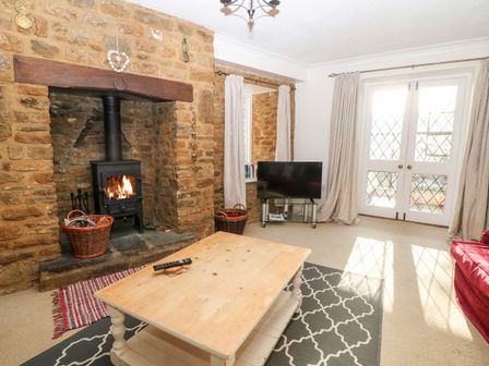A living room with a stone fireplace and wood burning stove a wooden coffee table a television on a stand and French doors with curtains at Stone Wheel Cottage in Hook Norton