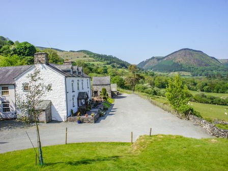 A white stone house with a gravel driveway and green hills in the background at Pengwern in Llangynog
