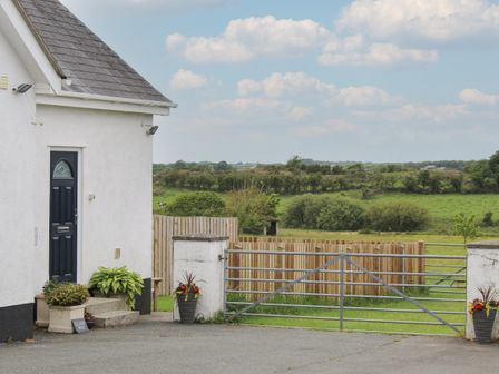 Side view of a white house with a black door near a metal gate and wooden fence overlooking green fields at Llain Mai in Llangefni