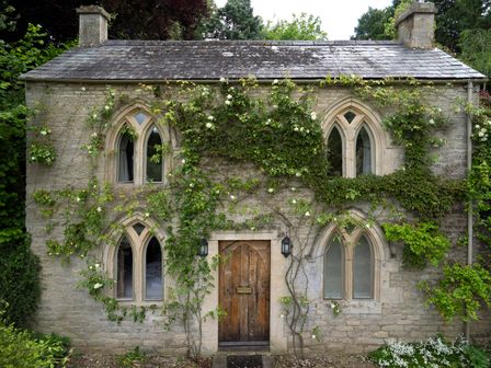 Stone house with arched windows and wooden door covered in climbing plants at All Souls Cottage Eastleach Cotswolds