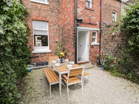 An outdoor patio area with a wooden dining table and benches set with dishes and blue cups next to a red brick house with plants and flowers at West Cliff House in Whitby