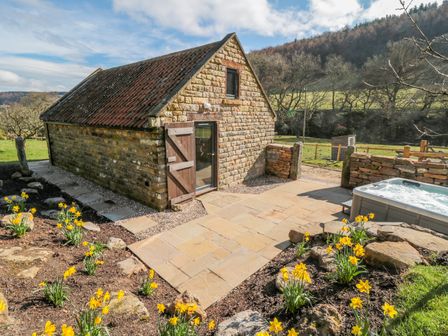 A stone building with a tiled roof next to a patio with yellow flowers and a hot tub at Thompson Rigg Barn in Langdale End near Thornton Dale