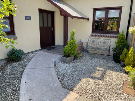 Entrance to a cottage with a curved concrete path a wooden door a metal bench and plants at Stable Cottage in Shipham
