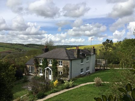 A stone and white two-story house with chimneys and a front door surrounded by greenery and hills in the background at Ribby Farmhouse in Lerryn