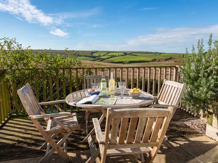 An outdoor wooden table with chairs on a deck with bottles and glasses overlooking green hills at 164 Cumber Close in Malborough