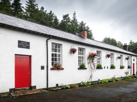A white building with red doors and windows with flower boxes and hanging flower baskets surrounded by trees at The Old School in Gortin