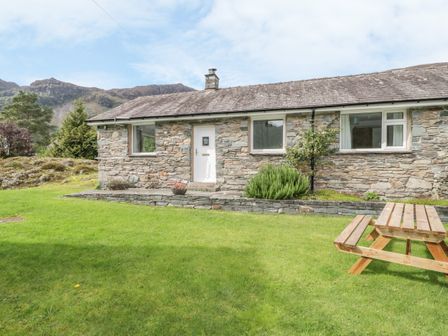 A stone cottage with a white door and windows on a grassy lawn with a wooden picnic table and hills in the background at Rock Cottage in Keswick