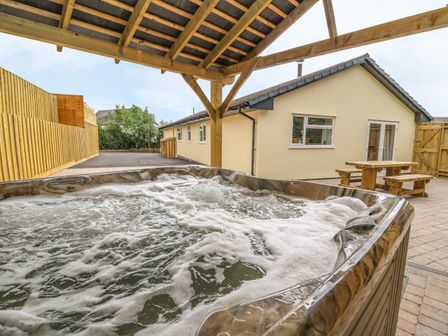 An outdoor hot tub under a wooden pergola with a yellow house and wooden picnic table in the background at New Peny in Llandrindod Wells