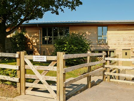 A wooden fenced entrance with gates and a cottage with wooden paneling in the background at Oak Apple in Teigngrace near Newton Abbot
