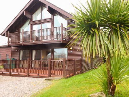 A two-story wooden house with balconies and a fenced porch with a tree and grass in the foreground at Lodge Sixty Six in St Columb Major