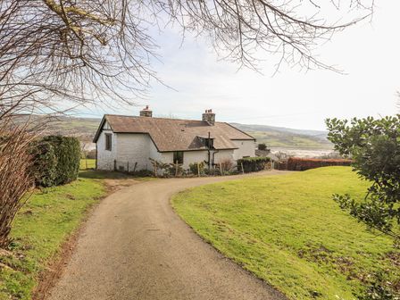 A single-story white cottage with a tiled roof at the end of a curved driveway with grass and trees around at Cymryd Uchaf in Conwy