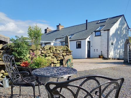 An outdoor seating area with a metal table and chairs near a stone wall and a white house at La'al Riddings Lowick Green Riddings Farm Near Ulverston