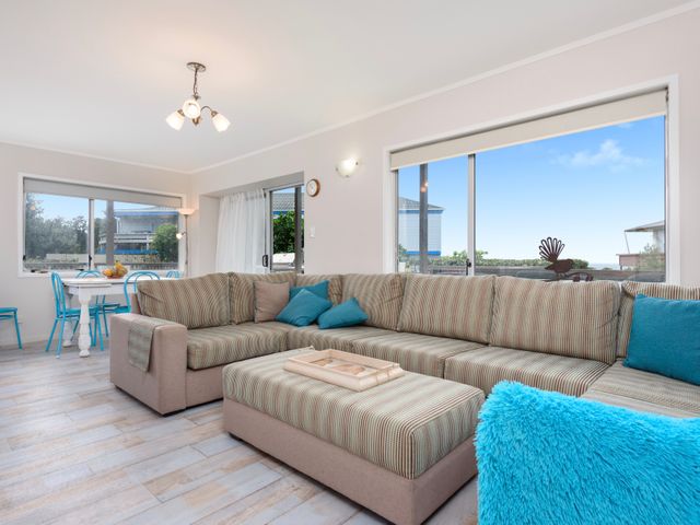 A living room with a striped sectional sofa and blue cushions next to large windows with a view at Laze on Loop - Waihi Beach Holiday Home in Waihi Beach