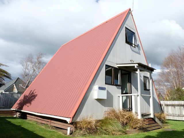 An A-frame house with a red roof and small porch surrounded by grass and shrubs at Central Ski Chalet Ohakune