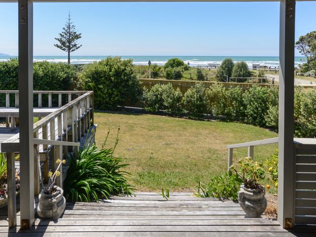 A wooden deck with steps leading to a grassy yard bordered by bushes and a view of the ocean with waves and a tree at Bach 112 - Waimarama Holiday Home in Waimarama