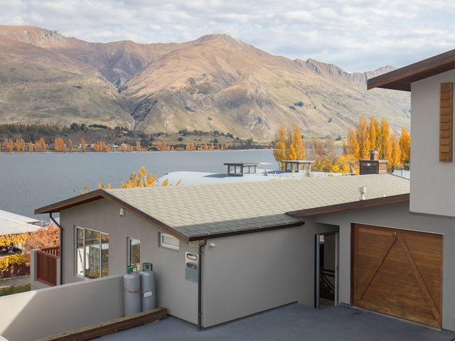A house with a wooden garage door overlooking a lake with mountains and trees in the background at Lakeview Heights Wanaka Home Bachcare NZ in Wanaka