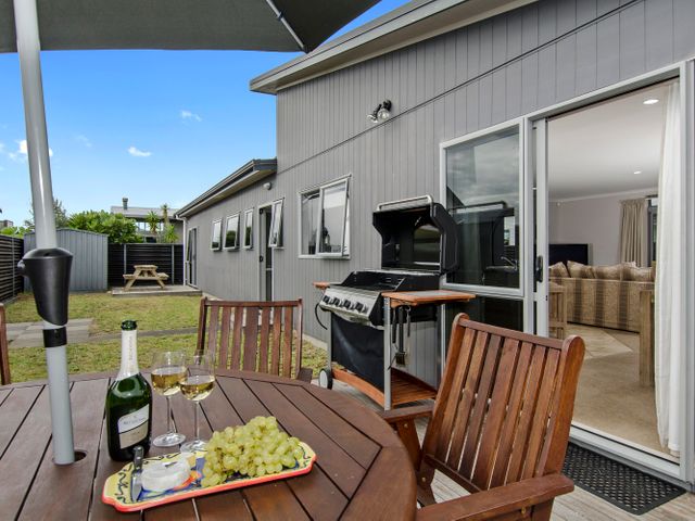 A patio with a wooden table and chairs a bottle of wine and grapes on the table a barbecue grill and an open sliding door showing a living room at Beauty on Bream Bay - Ruakaka Holiday Home in Ruakaka