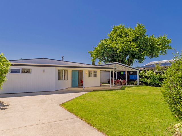 A white house with a blue door a concrete driveway and a green lawn at Gone Coastal - Whangamata Holiday House in Whangamata