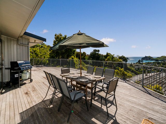 An outdoor wooden deck with a dining table set and umbrella a barbecue grill and coastal view at At Cathedral Cove - Hahei Holiday Home Hahei