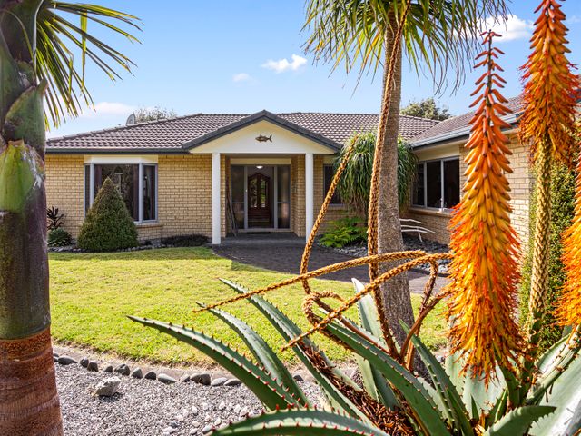 The front view of a single story brick house with a tiled roof and a garden with various plants and flowers at Fantails Roost - Bowentown Holiday Home Waihi Beach