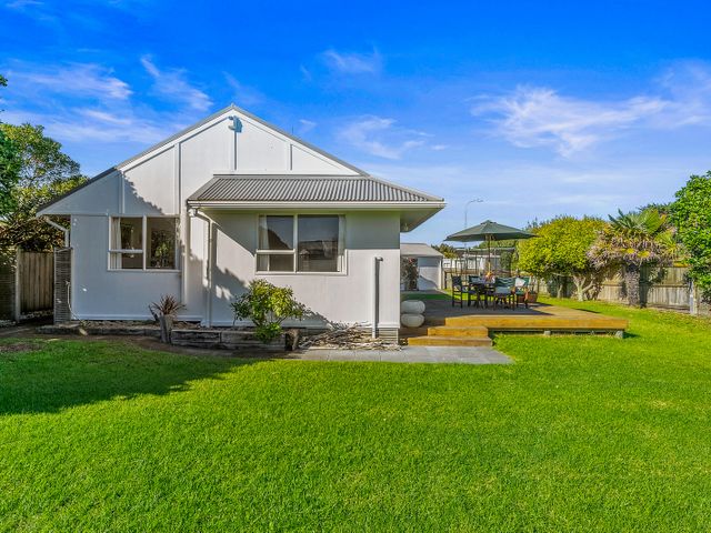 A backyard with green grass a wooden deck with table chairs and umbrella near a white house at Babbacombe Bach - Otaki Beach Holiday Home in Otaki Beach
