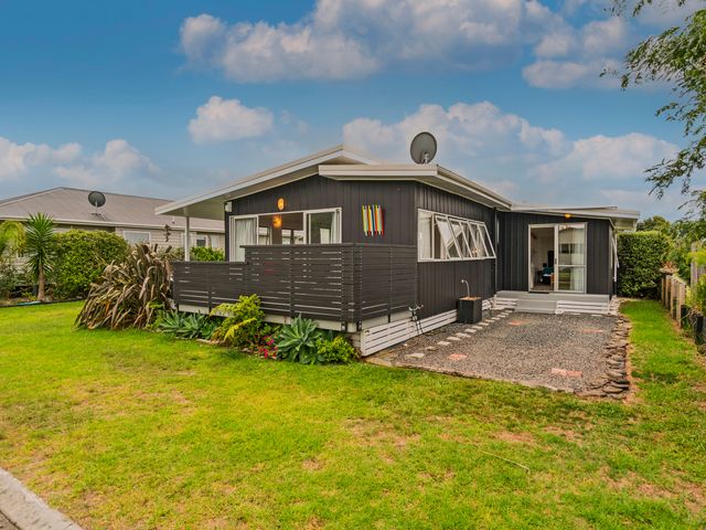 A black wooden house with a fenced porch and a gravel pathway surrounded by grass at Bach Life on Seabreeze - Whangamata Holiday Home Whangamata