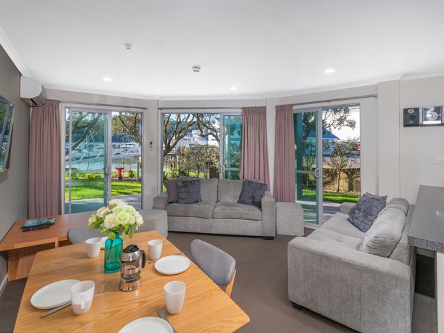 A living area with two sofas a wooden dining table set with plates and cups and sliding glass doors showing a garden and water view at Marinaview Whitianga Holiday Apartment Whitianga