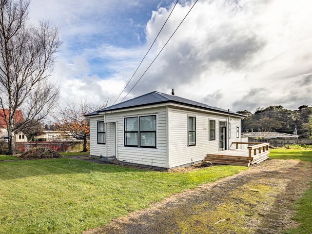 A small white house with multiple windows and a wooden deck surrounded by grass and trees at Rata Retreat - Ohakune Holiday Home in Ohakune