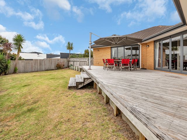 A wooden deck with a table and red chairs under an umbrella in a backyard with grass and palm trees at Paradise at the Bay - Ruakaka Holiday Home in Ruakaka