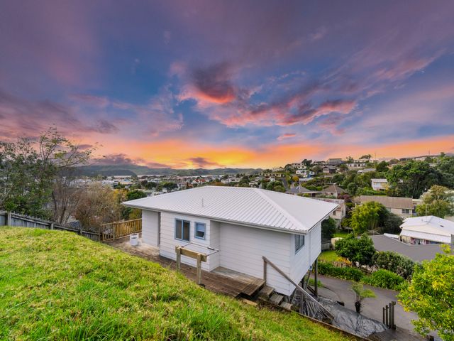 A house on a hillside with a wooden deck and surrounding neighborhood under a colorful sky at sunset at Raglan Beach Retreat in Raglan