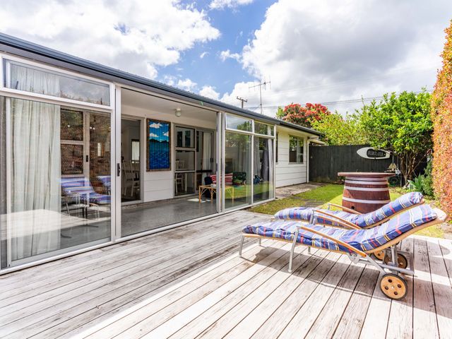 An outdoor wooden deck with two lounge chairs and a barrel table next to a house with sliding glass doors at Ruakaka Holiday Escape Ruakaka