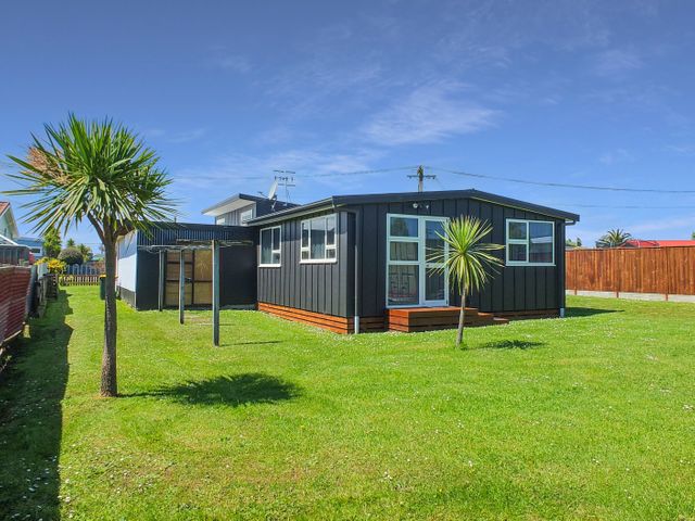 A single story dark exterior house with a green lawn and two small trees in the yard at Snapper Bach - Foxton Beach Holiday Home in Foxton