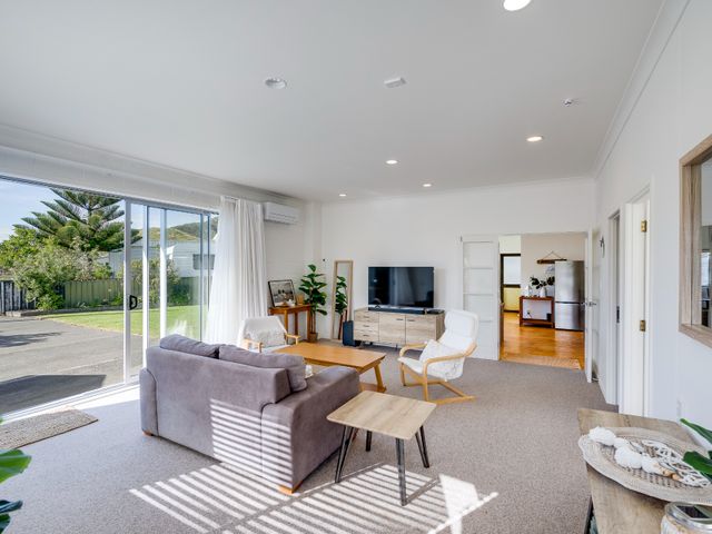 A living room with a sofa, two chairs, a coffee table, a television on a wooden stand, and sliding glass doors at Mahia Bliss - Surf & Serenity Holiday Home Mahia Peninsula