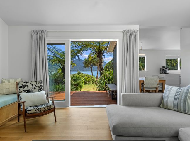 A living room with a sliding glass door opening to a wooden deck and garden with trees and water view at Luxury Waterfront Retreat at Mahau Sound in Mahau Sound
