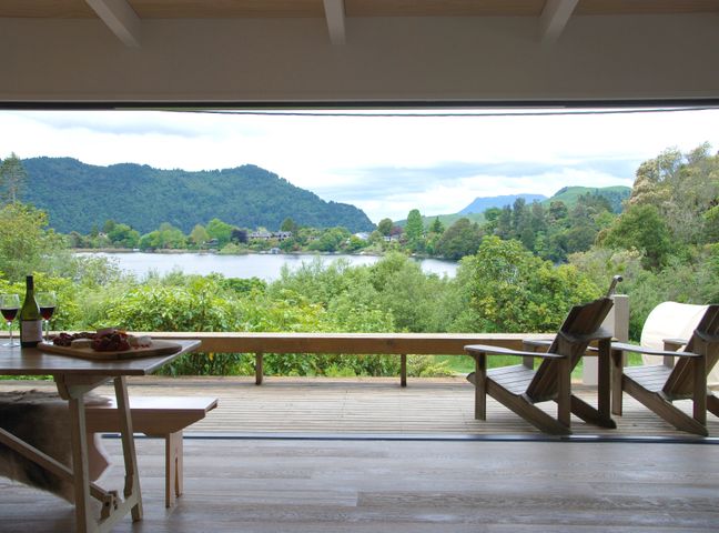 A deck with two wooden chairs and a table with wine and snacks overlooking a lake and mountains at Lake View Okareka - Lake Okareka Holiday Home