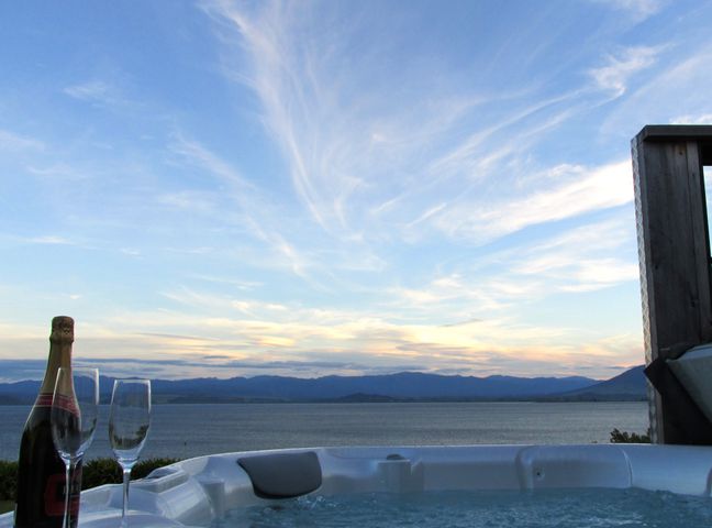 A hot tub with a bottle of champagne and two champagne flutes overlooking a body of water and mountains at Waipapa Cottage - Kuratau Holiday House in Kuratau