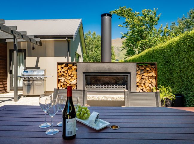 An outdoor patio with a wooden table set with wine glasses a wine bottle and grapes a wood storage fireplace and a barbecue grill at Modern Lake Haven Wanaka Holiday Home Wanaka
